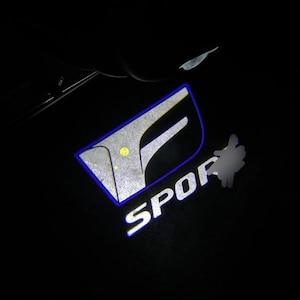 Pode incluir: Logotipo iluminado da porta do carro com a palavra "SPORT" em branco. O logotipo apresenta um design "F" estilizado com uma contorno azul e um interior prateado, e um ponto amarelo.