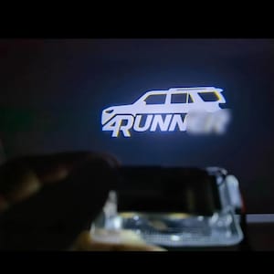 May include: A white illuminated projection of a vehicle with the word "4Runner" in a stylized font. The image is projected onto a dark surface, with the vehicle's outline clearly visible. The overall effect is a bright, eye-catching display.