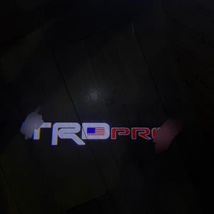 Puede incluir: Logotipo TRD PRO iluminado en blanco, rojo y azul, con un diseño de bandera estadounidense. El logotipo se proyecta sobre una superficie de madera oscura, creando un efecto visual llamativo.
