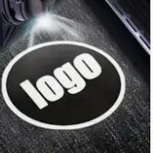 Peut inclure: Un autocollant ovale noir avec le mot "logo" en blanc, éclairé par une lampe de poche. L'autocollant a une bordure blanche et est placé sur une surface sombre et texturée. L'image est un gros plan.