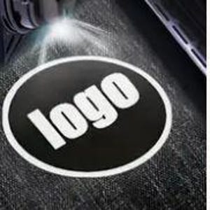 Puede incluir: Una pegatina ovalada negra con la palabra "logo" en blanco, iluminada por una linterna. La pegatina tiene un borde blanco y está colocada sobre una superficie oscura y texturizada. La imagen también muestra una parte de un objeto negro.
