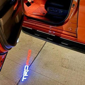 May include: An open orange car door with a black running board. The illuminated text "TRD PRO" is projected onto the ground in white and orange. The interior of the car is visible.
