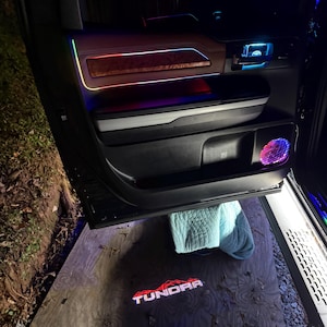Peut inclure: Intérieur de porte de voiture avec éclairage d'ambiance multicolore. Le panneau de porte présente des accents de grain de bois et une finition noire. La porte ouverte projette une lumière au sol avec le mot "TUNDRA". L'éclairage comprend des teintes rouges, bleues et vertes.