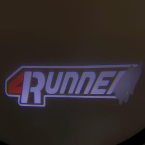 Peut inclure: Panneau lumineux avec le texte "4Runner" dans une police stylisée. Le "4" est rouge, et le mot "Runner" est blanc avec un contour bleu. Le panneau rectangulaire semble être un accessoire de voiture.