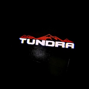以下が含まれることがあります： 黒い背景に、白い文字で「TUNDRA」と書かれ、その上に赤い山脈のデザインが施されています。この画像はロゴまたはエンブレムです。