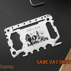 Puede incluir: Una tarjeta multiherramienta plateada con varios recortes y una regla a lo largo del borde superior. La tarjeta presenta una escena de campamento con el texto "William's Family". También son visibles las palabras "Bottle Opener" y "Protractor".