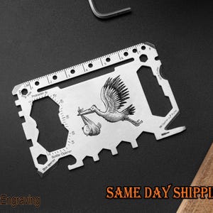 May include: A silver multi-tool with a black and white engraved image of a stork carrying a baby. The tool features a ruler, bottle opener, box opener, and other functions. The text "SAME DAY SHIPPING" is visible in the lower right corner.
