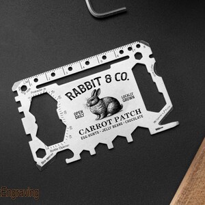 Puede incluir: Una tarjeta multiherramienta plateada con el texto "RABBIT & CO." y una ilustración de un conejo. Incluye una regla, un transportador, un abrebotellas y un abridor de cajas. La tarjeta también tiene el texto "CARROT PATCH" y "LOCALLY GROWN".