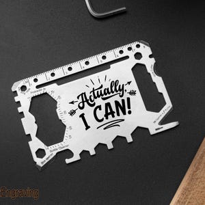 Könnte beinhalten: Eine silberne Multi-Tool-Karte mit dem Text "Actually, I CAN!" und verschiedenen Aussparungen. Sie enthält ein Lineal mit Zentimetermarkierungen und einen Flaschenöffner. Die Karte liegt auf einer dunklen Oberfläche.