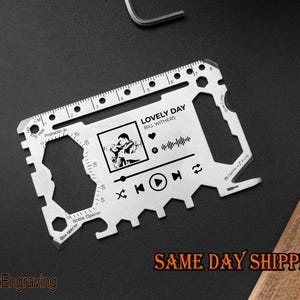 May include: A silver multi-tool card with various cutouts and functions, including a ruler, bottle opener, and protractor. The card features a music player interface with the text "LOVELY DAY" by Bill Withers and a graphic of a person. The text "FranzEngraving" is also visible.