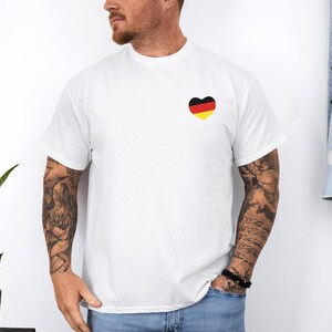 May include: White t-shirt with a black, yellow, and red heart-shaped design on the left chest. The heart design resembles the German flag.