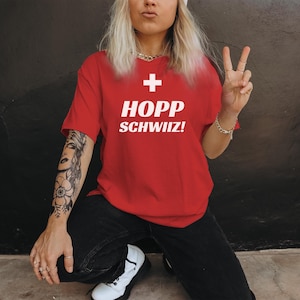 May include: A red t-shirt with a white Swiss cross and the text "HOPP SCHWIIZ!" printed on the front. The shirt is being worn by a person with a tattoo on their arm.