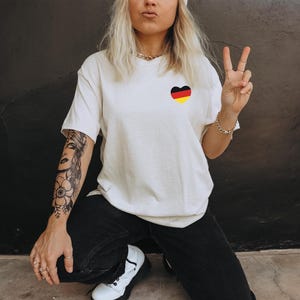 May include: A white t-shirt with a black and red heart design. The heart is in the shape of the German flag. The person wearing the shirt has a black and white tattoo on their arm.