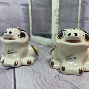 May include: Two ceramic frog figurines with brown and yellow painted details. The frogs are sitting with their mouths open and have black eyes. The bottom of each frog has the text "La  Parrilla" written on it.