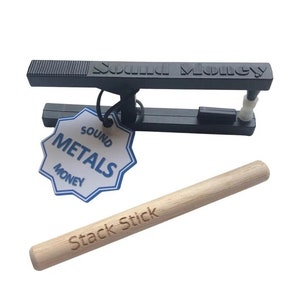 May include: A black metal Sound Money device with a tag that reads "Sound Metals Money." A light-colored wooden "Stack Stick" is also present. The device has a textured top and a white plastic component.