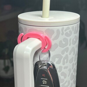 May include: White travel mug with a leopard print design and a pink key holder attached to the handle. A car key is attached to the key holder. A white straw is in the lid of the mug.