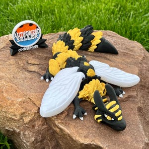 May include: A detailed, handcrafted dragon figurine with yellow and black segments, white wings, and black claws. The dragon is posed on a rock, with an "AUTHORIZED SELLER CINDERWING" logo nearby. The background is green grass.