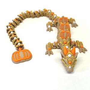 May include: A whimsical, articulated dragon figurine in shades of tan, orange, and gold. The dragon's body is segmented, with pumpkin-shaped accents and white and orange details. The head features intricate gold detailing and orange accents.