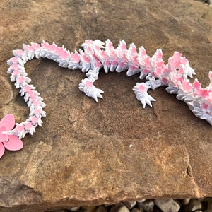 May include: A pink and white origami dragon made of paper. The dragon is lying on a brown rock.