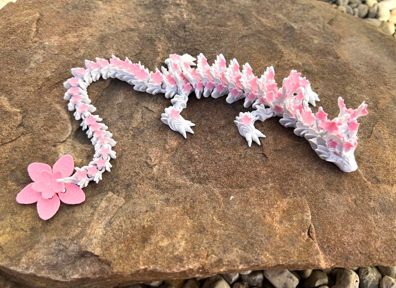 May include: A pink and white origami dragon made of paper. The dragon is lying on a brown rock.