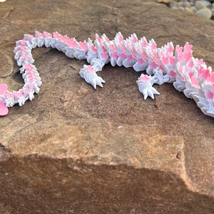 May include: A pink and white 3D printed dragon figurine with a flower tail. The dragon is lying on a brown rock.