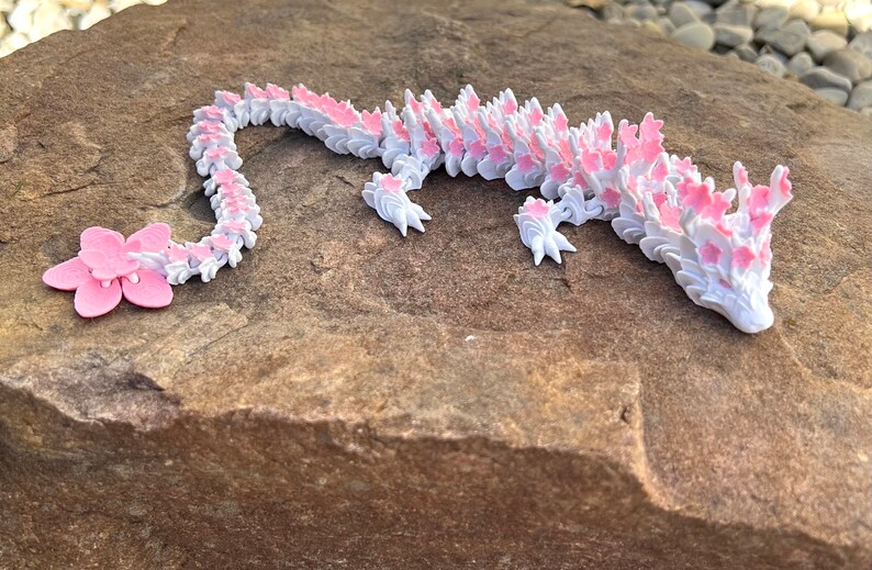 May include: A pink and white 3D printed dragon figurine with a flower tail. The dragon is lying on a brown rock.