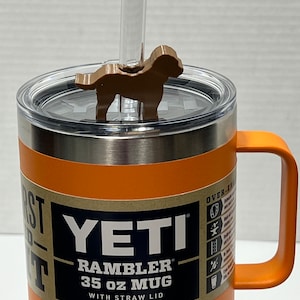 May include: Orange Yeti Rambler 35 oz mug with a straw lid and a brown dog-shaped lid topper. The mug has a stainless steel exterior and a brown label that reads "YETI Rambler 35 oz Mug with Straw Lid".