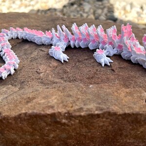 May include: A white and pink origami dragon made of paper. The dragon is lying on a brown rock.