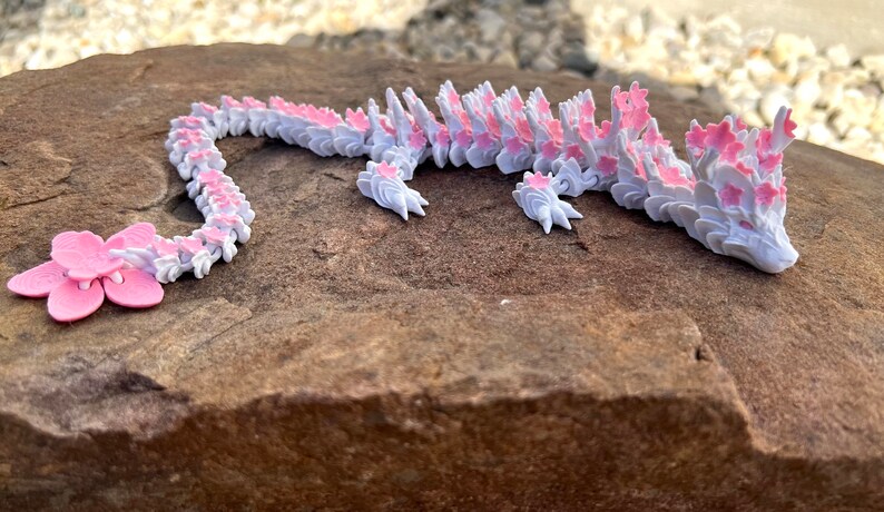 May include: A white and pink origami dragon made of paper. The dragon is lying on a brown rock.