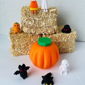 Pode incluir: Uma coleção de estatuetas com tema de Halloween, incluindo uma abóbora, um fantasma, um milho doce, um esqueleto e dois morcegos. As estatuetas estão dispostas em um leito de palha.