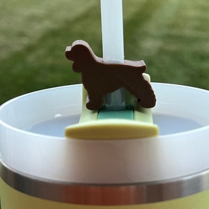 Puede incluir: Una taza de viaje amarilla y blanca con un soporte para pajitas en forma de perro marrón. El soporte para pajitas está unido a la tapa de la taza y tiene un diseño verde y blanco.