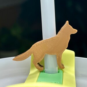 May include: A brown plastic wolf silhouette attached to a clear plastic straw. The straw is inserted into a yellow plastic lid with a green plastic insert.