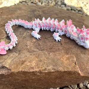 May include: A white and pink origami dragon with a long tail and a flower design. The dragon is lying on a brown rock.