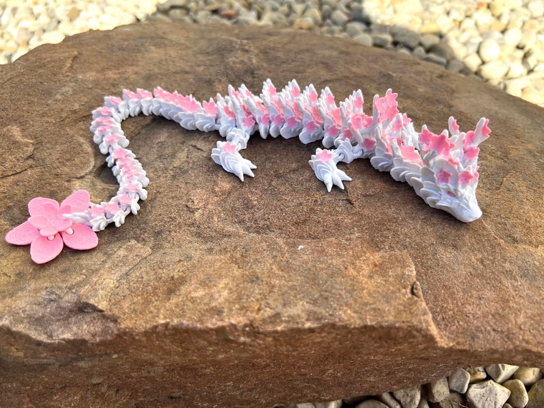 May include: A white and pink origami dragon with a long tail and a flower design. The dragon is lying on a brown rock.