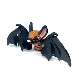 May include: A handcrafted bat figurine with a brown head, large black wings, and big eyes. The bat has pink inner ears and a small pink nose. The wings are spread, and the bat appears to be in flight. The figurine is set against a white background.