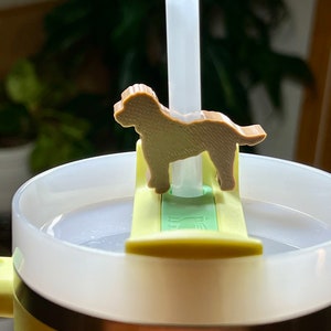 Może przedstawiać: Żółty kubek podróżny z brązowym uchwytem na słomkę w kształcie psa i przezroczystą słomką z tworzywa sztucznego. Uchwyt na słomkę jest przymocowany do pokrywki kubka.