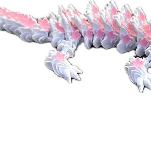 May include: A white and pink 3D printed dragon with a long tail and a flower at the end. The dragon is made up of many small, interlocking pieces.