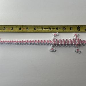 May include: A pink and white paper chain in the shape of a lizard, measuring approximately 30 cm long. The chain is made of individual paper links, each with a pink flower design.