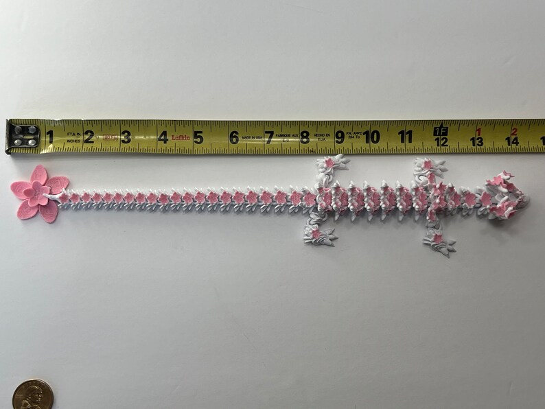 May include: A pink and white paper chain in the shape of a lizard, measuring approximately 30 cm long. The chain is made of individual paper links, each with a pink flower design.