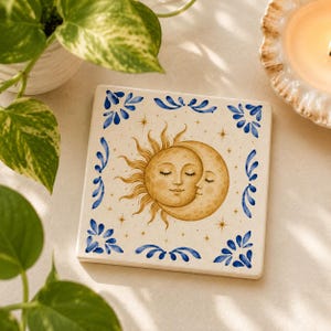 May include: A square ceramic coaster featuring a central illustration of a golden sun and crescent moon with closed eyes. The design is surrounded by a blue floral border pattern and small gold stars on a cream-colored background.