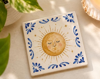 May include: A square ceramic tile featuring a hand-painted golden sun with a serene face and closed eyes at the center. The design includes radiating golden sunbeams, small golden stars, and a decorative blue floral border pattern.