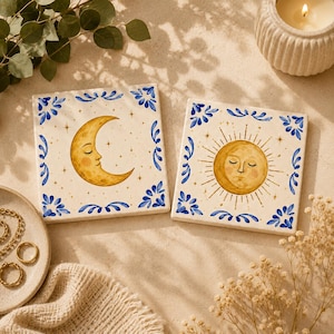 May include: Two square ceramic tiles featuring hand-painted celestial designs. One tile displays a golden crescent moon with a face, while the other shows a golden sun with a face. Both tiles are bordered by blue floral patterns on a cream background.