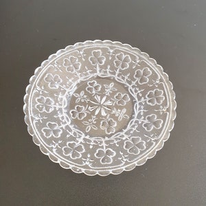 May include: A clear glass plate featuring a scalloped edge and an embossed pattern of shamrocks and vines. The centre of the plate displays a star-like geometric design surrounded by a ring of clover motifs, all rendered in a transparent finish.
