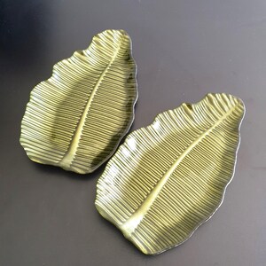 May include: Two metallic olive green decorative trays shaped like elongated tropical leaves. Each tray features a central vein and detailed ribbed texture radiating outwards, mimicking the natural appearance of a leaf with a glossy finish.