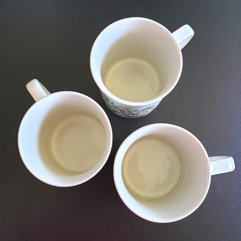 May include: Three white ceramic mugs with handles are arranged on a dark surface. The mugs have a simple design, with a light-colored interior and a white exterior. One mug has a floral pattern.
