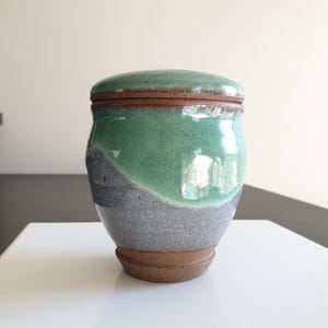 May include: A ceramic jar with a lid, featuring a glossy mint green glaze on the upper portion and a textured, matte grey finish on the lower section. The base and the rim are made of exposed, unglazed brown clay, creating a distinct contrast.