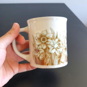 May include: Cream-colored ceramic mug with a floral design featuring daffodils in shades of brown and light blue. The mug has a curved handle and a slightly flared rim. The floral pattern is embossed on the mug's surface.