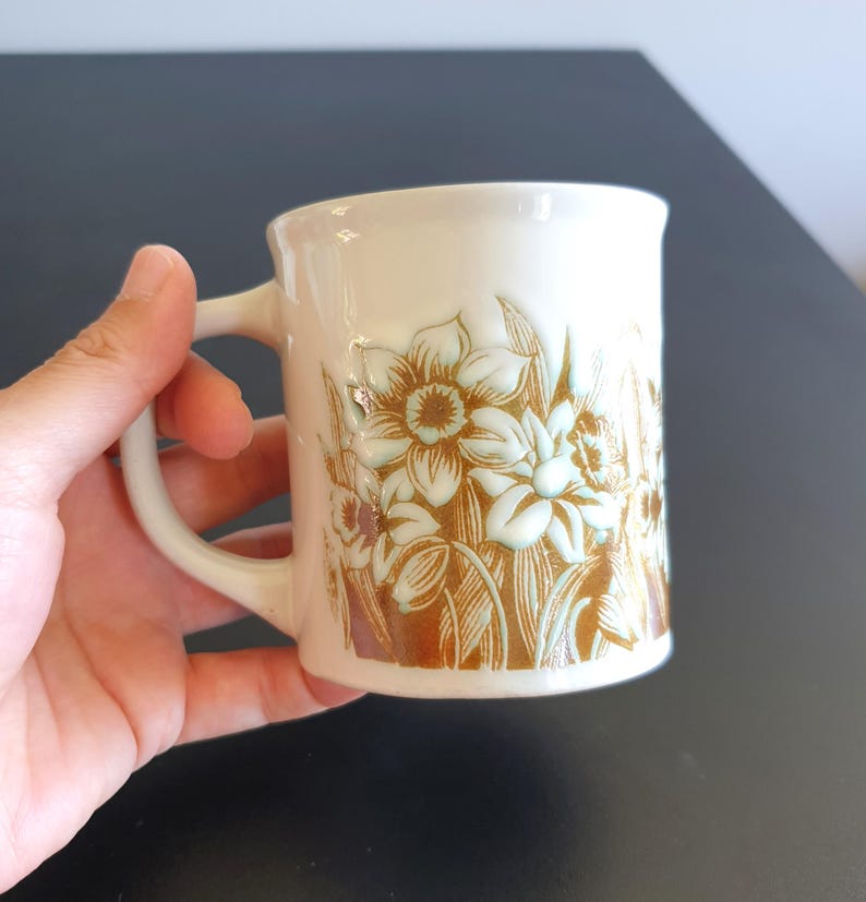 May include: Cream-colored ceramic mug with a floral design featuring daffodils in shades of brown and light blue. The mug has a curved handle and a slightly flared rim. The floral pattern is embossed on the mug's surface.
