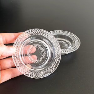 May include: Two clear glass coasters with a beaded edge design. The coasters are round and have a slightly raised centre.