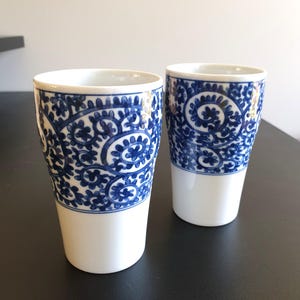 May include: Two white ceramic tumblers with a blue paisley design on the upper half. The tumblers have a simple, elegant shape and are suitable for serving beverages. The blue pattern contrasts with the white base.